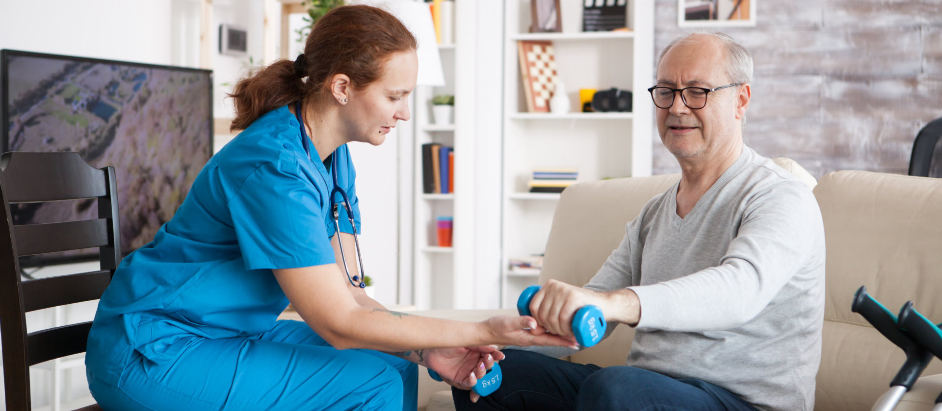  Female nurse doing physiotherapy with senior man in nursing home using dumbbells.
